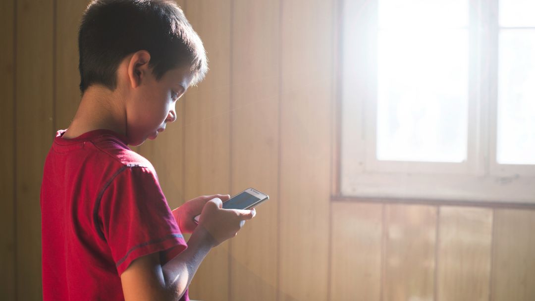 A young child alone by a window looking at a phone